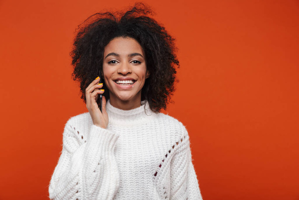 Attractive young african woman wearing sweater talking on mobile phone isolated over red background - Foto, afbeelding