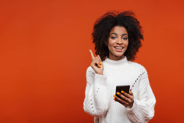 Attrayant sourire jeune femme africaine portant pull tenant téléphone portable isolé sur fond rouge, pointant du doigt
 - Photo, image