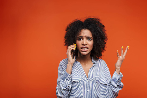 Angry young african woman talking on mobile phone isolated over red background - Foto, afbeelding