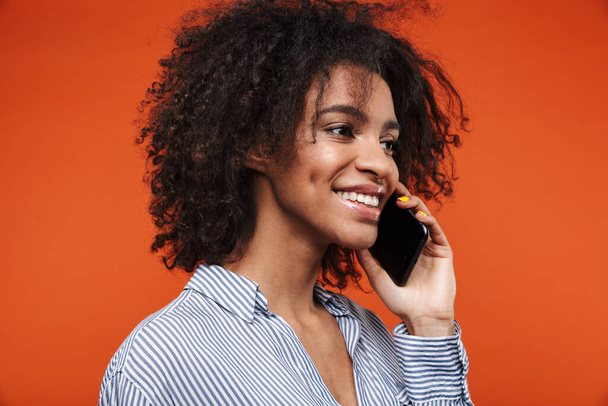Gülümseyen çekici Afrikalı kadın kırmızı arka planda izole bir şekilde cep telefonuyla konuşuyor. - Fotoğraf, Görsel
