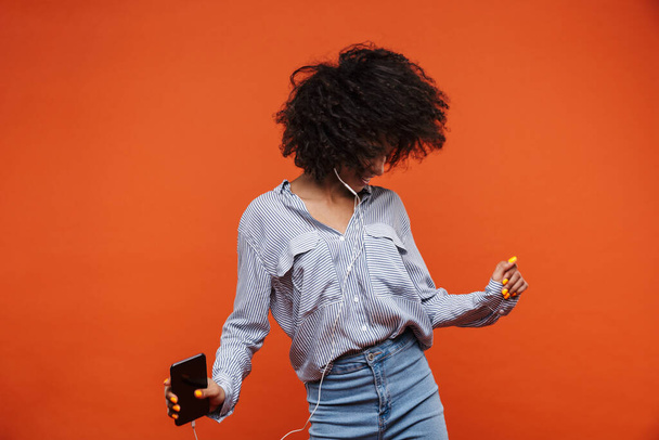 Smiling young beautiful african woman wearing casual clothes standing isolated over red background, listening to music with earphones and mobile phone, dancing - Fotografie, Obrázek