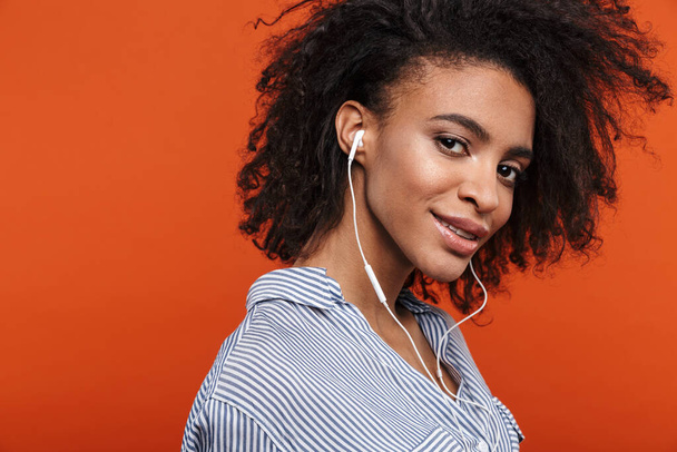 Smiling young beautiful african woman wearing casual clothes standing isolated over red background, listening to music with earphones - Foto, Bild