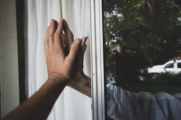Eine ältere Frau hält von zu Hause aus eine Hand ans Fenster und berührt die Hände ihres Enkels, der während der Quarantäne wegen der Coronavirus-Pandemie von außen zu Besuch ist - Foto, Bild