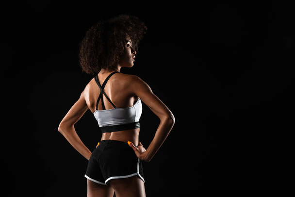 Image from back of african american woman in sportswear posing with hands on her waist isolated over black background - Foto, imagen