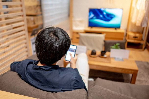 Νεαρός Ασιάτης με casual ρούχα κάθεται σε άνετο καναπέ (καναπέ) χρησιμοποιώντας smartphone και βλέποντας τηλεόραση στο σαλόνι. Άνετος τρόπος ζωής στο σπίτι. Χαλαρωτική έννοια της οικιακής ζωής - Φωτογραφία, εικόνα