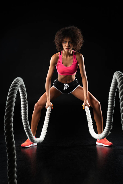 Image of athletic african american sportswoman doing exercise with battle ropes isolated over black background - Valokuva, kuva