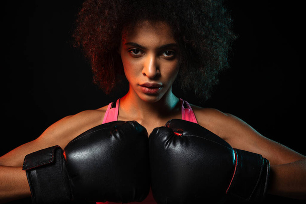 Image of young african american sportswoman working out in boxing gloves isolated over black background - Фото, изображение