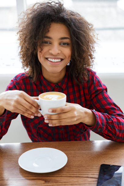 Portrait of pretty young african american woman wearing plaid shirt smiling and drinking coffee in cafe - Valokuva, kuva