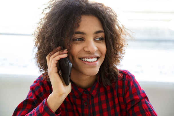Portrait of pretty young african american woman wearing plaid shirt drinking coffee and talking on cellphone in cafe - Photo, image
