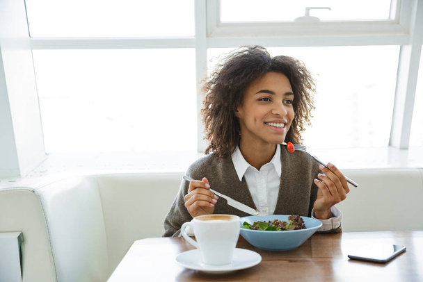 Portrait of young beautiful african american woman eating salad while having lunch in cafe - Фото, изображение