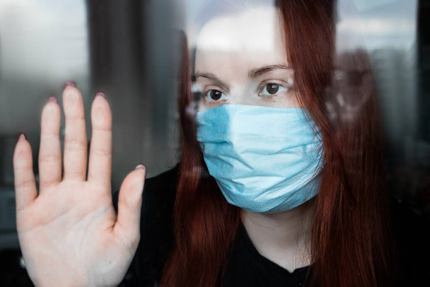 Μια θλιμμένη νεαρή γυναίκα με ιατρική μάσκα κοιτάζει έξω από το παράθυρο. Καραντίνα έννοια κατά τη διάρκεια της πανδημίας coronavirus. - Φωτογραφία, εικόνα