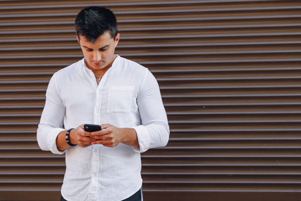 young stylish guy in shirt typing on phone on simple brown background - Photo, Image