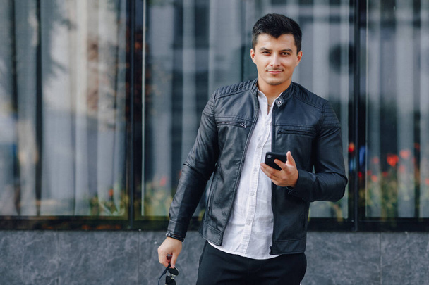 young stylish guy in glasses in black leather jacket with phone on simple glass background - Foto, imagen