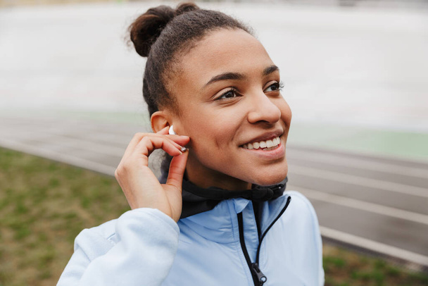 Attractive smiling young healthy african sportswoman listening to music with wireless earphones wrking out at the stadium - Фото, изображение