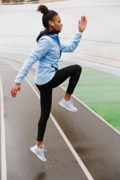 Beautiful young fit african sportswoman wearing wireless earphones doing stretching exercises at the stadium, jumping - Photo, Image
