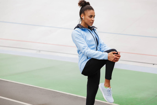 Beautiful young fit african sportswoman wearing wireless earphones doing stretching exercises at the stadium - Photo, Image