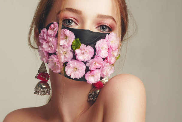 Mode-Foto von einem Mädchen in einer Maske aus Blumen. Frühling, den wir nicht atmen können. Virus, Pandemie, Coronavirus, Maskenmodell, Schönheitsmodel in einer Maske aus frischen rosa Blüten. Mädchen mit zartem rosa Make-up. - Foto, Bild