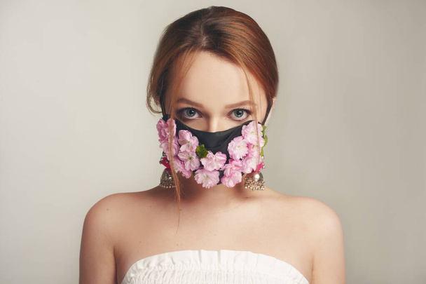 花のマスクに女の子のファッション写真。呼吸できない春。ウイルス、パンデミック、コロナウイルス、マスクモデル、新鮮なピンクの花のマスクの美しさモデル。繊細なピンクの化粧をした女の子. - 写真・画像