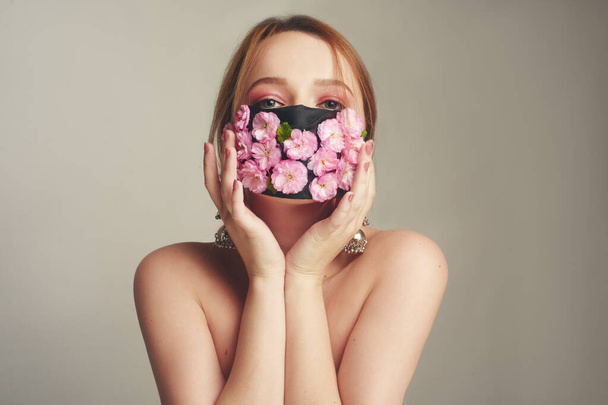 Mode-Foto von einem Mädchen in einer Maske aus Blumen. Frühling, den wir nicht atmen können. Virus, Pandemie, Coronavirus, Maskenmodell, Schönheitsmodel in einer Maske aus frischen rosa Blüten. Mädchen mit zartem rosa Make-up. - Foto, Bild