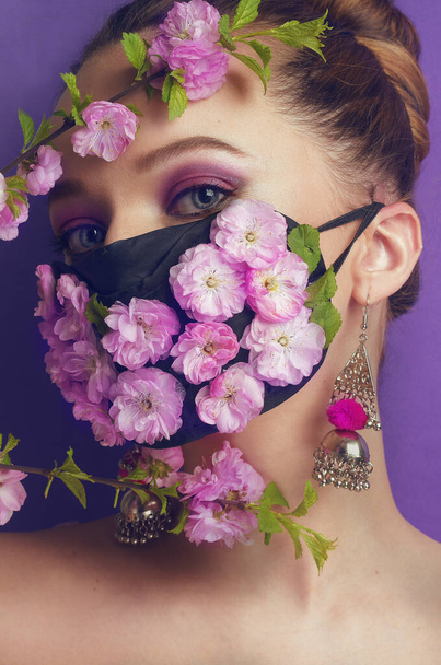 Prachtig lentemeisje, met bloemen op een masker. Lente die we niet kunnen ademen, de geur van bloemen. Portet van een meisje op een paarse achtergrond in een masker van bloesems en bloemknoppen. Coronavirus, maskers, pandemie. - Foto, afbeelding
