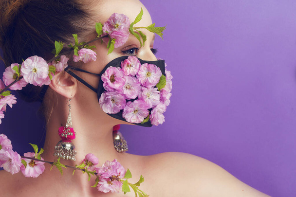 Πανέμορφο ανοιξιάτικο κορίτσι, με λουλούδια στη μάσκα. Άνοιξη που δεν μπορούμε να αναπνεύσουμε, η μυρωδιά των λουλουδιών. Πόρτα ενός κοριτσιού σε μωβ φόντο με μια μάσκα από άνθη και μπουμπούκια λουλουδιών. Ιός του Coronavirus, μάσκες, πανδημία. - Φωτογραφία, εικόνα