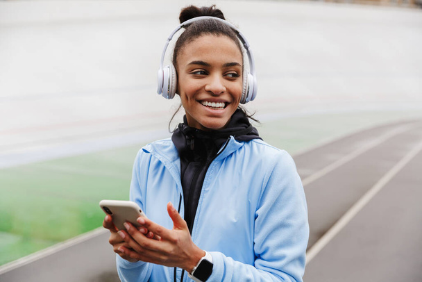 Ελκυστική χαμογελαστή νεαρή υγιής Αφρικανή αθλήτρια που ακούει μουσική με ασύρματα ακουστικά να βγαίνουν στο γήπεδο, κρατώντας κινητό τηλέφωνο - Φωτογραφία, εικόνα