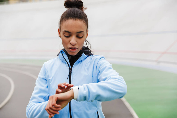 Attractive young african sportswoman resting after running at the stadium, looking at smartwatch - Photo, Image