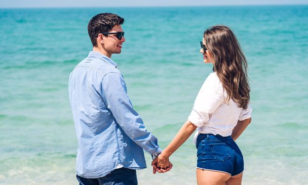 Διακοπές ρομαντικοί εραστές νεαρό ευτυχισμένο ζευγάρι αγκαλιά και στέκεται στην άμμο και ατενίζοντας τη θάλασσα διασκεδάζοντας και χαλαρώνοντας μαζί στην τροπική παραλία. - Φωτογραφία, εικόνα