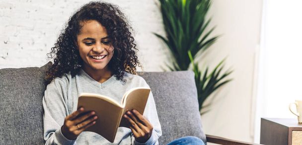 Joven hipster afroamericano mujer negra relajante leyendo el estudio del libro de papel y mirando la revista de la página mientras está sentado en el sofá en casa
 - Foto, Imagen