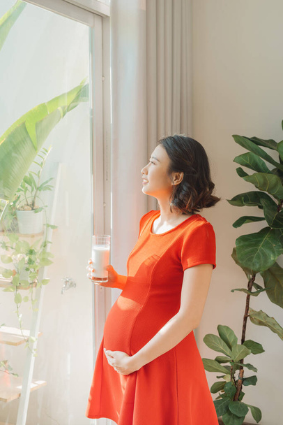 Young Asian pregnant woman holding milk glass when standing behind the window. - Photo, image
