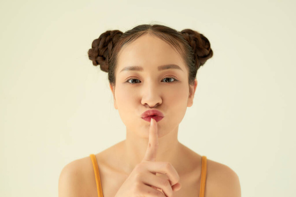 Držte se v tajnosti! Portrét hravý pozitivní asijské dospívající dívka s buchty účes ukazující ticho gesto přes světlo pozadí. - Fotografie, Obrázek