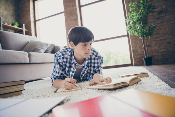 Closeup photo of teenager school boy lying floor near couch stay home quarantine time writing doing homework diligent student read book literature object living room indoors - Foto, imagen
