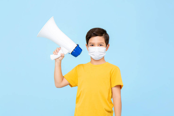 Симпатичний молодий хлопчик в медичній масці тримає мегафон ізольовано на світло-блакитному фоні
 - Фото, зображення
