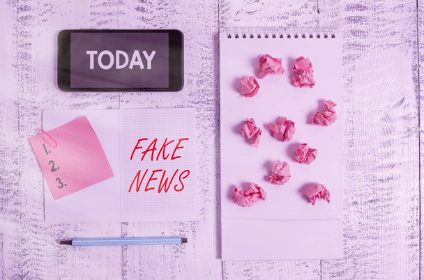 Word writing text Fake News. Business concept for Giving information to showing that is not true by the media Squared spiral notebook marker smartphone paper balls note clip wooden. - Photo, Image