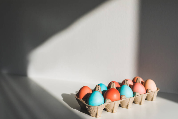 Scatola di cartone con uova di pollo blu e rosse dipinte per Pasqua. Tavolo bianco e bella luce del sole. Concetto pasquale
. - Foto, immagini