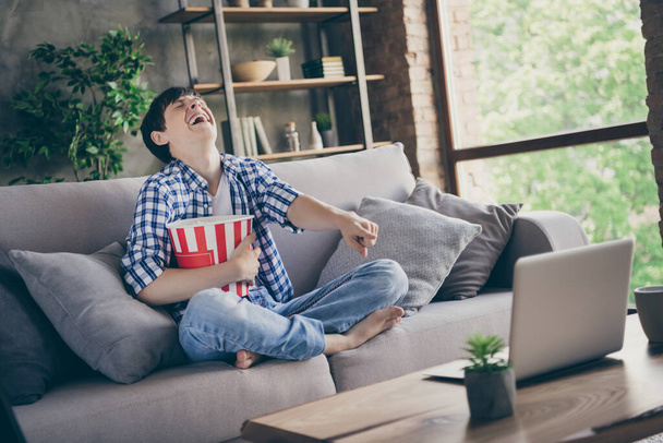 Photo of teen school boy sit comfy couch stay home quarantine time watch notebook movie laughing out loud direct finger screen eat popcorn good mood legs crossed living room indoors - Fotoğraf, Görsel