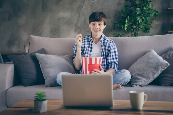 Photo of teen school boy sitting comfy couch stay home quarantine time watch notebook movie online show eat popcorn good mood legs crossed living room indoors - Zdjęcie, obraz