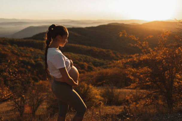 Young pregnant woman walking outdoors with amazing autumn mountain and forest view. Warm sunlight. Wellness and vitality in maternity time. - Photo, Image