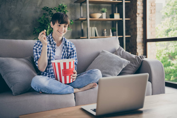Photo of teenager school boy sitting comfy couch stay home quarantine watch notebook movie online show eat popcorn good mood legs crossed living room indoors - Fotografie, Obrázek