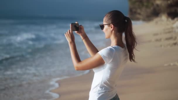 Cheerful young beautiful woman in sunglasses takes photos of beach - Materiał filmowy, wideo