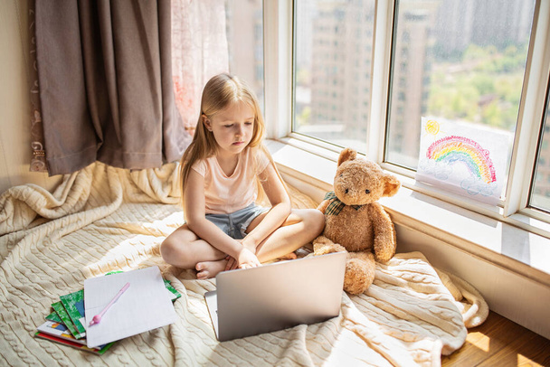 Pretty schoolgirl studying homework during her online lesson at home, social distance during quarantine covid-19, self-isolation, online education concept, home schooler. Workplace in living room - 写真・画像