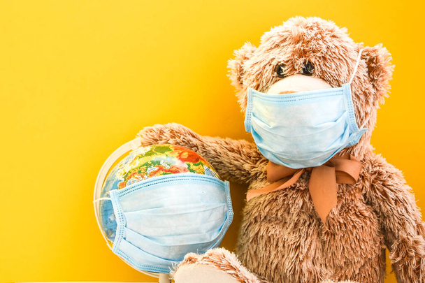 Teddybär-Spielzeug mit medizinischer Gesichtsmaske und Erdkugel mit Schutzmaske auf gelbem Hintergrund, Kopierplatz für Ihren Text, Quarantäne gegen Coronavirus, covid-19 - Foto, Bild