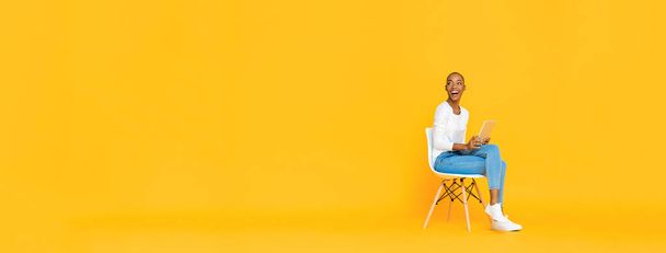 Trendy smiling African American woman sitting on a chair using tablet computer thinking and looking at empty space aside isolated yellow banner background - Fotografie, Obrázek