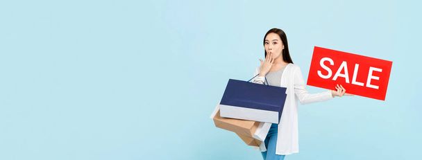 Sorpresa bella donna asiatica con borse della spesa che mostrano segno di vendita rosso isolato su sfondo banner blu chiaro con spazio copia
 - Foto, immagini
