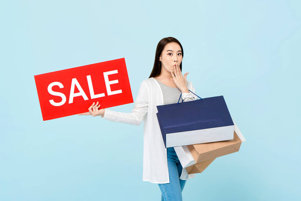 Surprised beautiful Asian woman with shopping bags showing red sale sign isolated on light blue background - Φωτογραφία, εικόνα