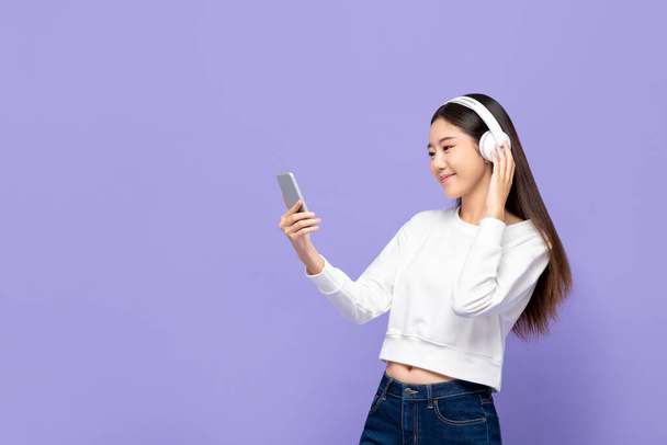Portrait of young smiling Asian woman listening to music on mobile phone using wireless headphone in purple isolated studio background - Photo, Image