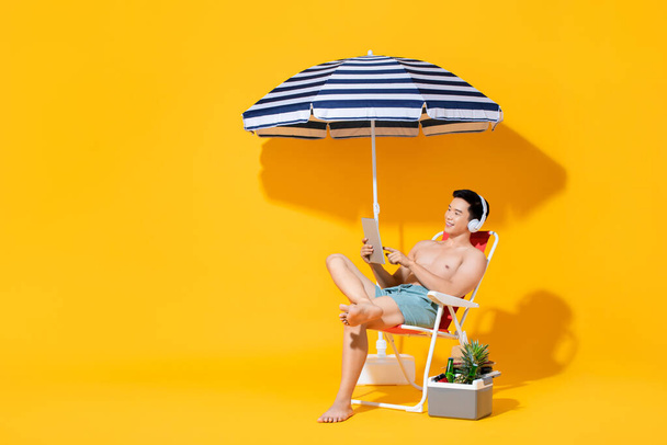 Retrato de un joven asiático sin camisa sentado en una silla de playa relajándose y escuchando música en un aislado fondo amarillo veraniego
 - Foto, imagen