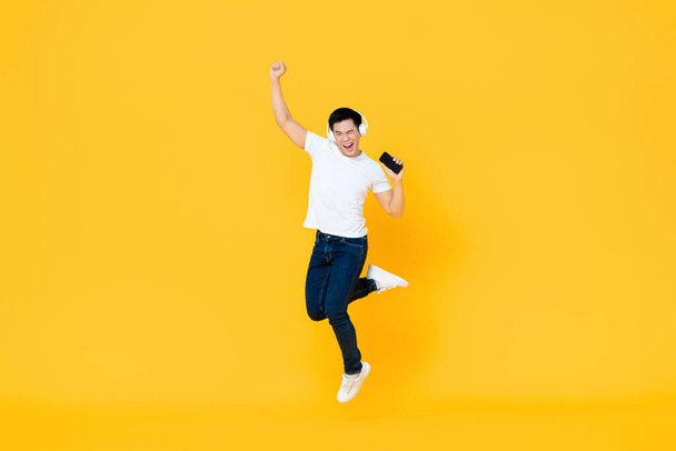 幸せな若いアジアの男は、携帯電話から音楽を聞いてヘッドフォンを身に着けていると黄色の背景に孤立調達拳でジャンプ - 写真・画像