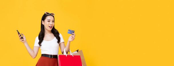 Young beautiful Asian woman making an online payment through mobile phone with credit card while carrying shopping bags in isolated yellow banner background with copy space - Photo, Image