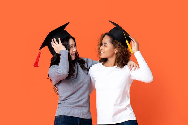 Retrato de dos mujeres felices de raza mixta graduándose sosteniendo allí gorras de graduación mirándose en el fondo del estudio aislado naranja
 - Foto, Imagen